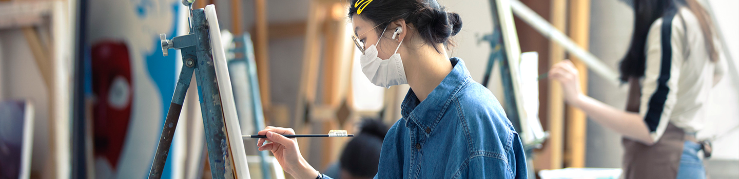 A person painting at an easel wearing a mask and a blue shirt.