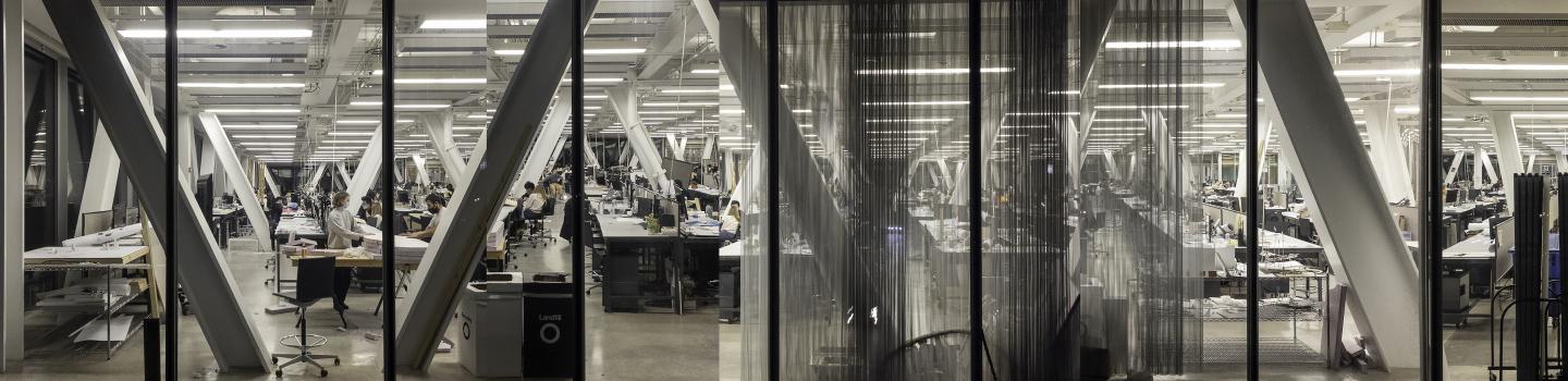 People working at desks with computer screens from inside a building with bright white lights and angled support beams.