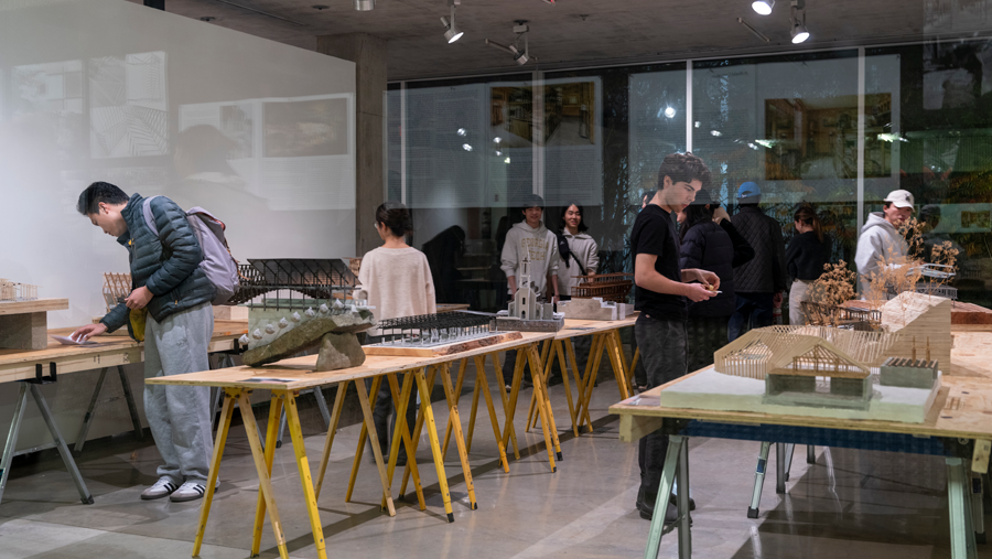 People gather around tables in a gallery covered with wooden and concrete architectural models
