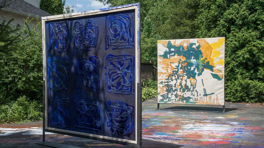 Two canvas paintings sit outside in the sun atop a multicolored painted pavement surface. The canvas in the foreground is royal blue and navy and the canvas in the background is teal, orange, yellow, and cream. 