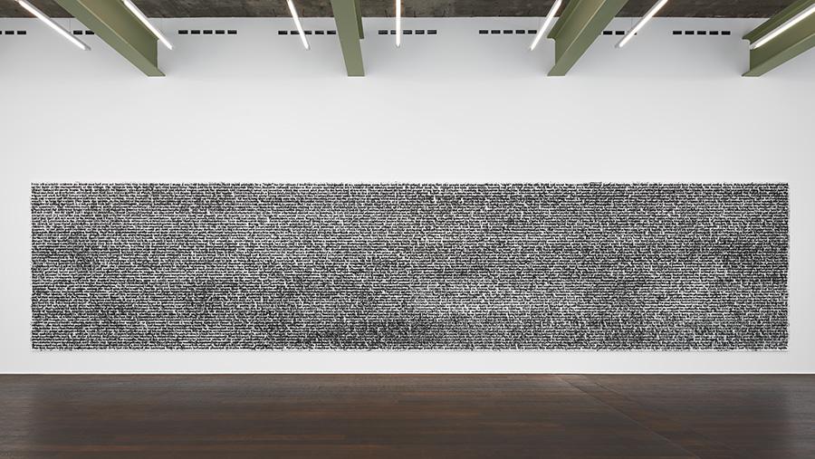 A photo of a white wall with a large, rectangular black painting on it. The painting looks like a long string of text, but the photo is taken from too far away to read any of the words.