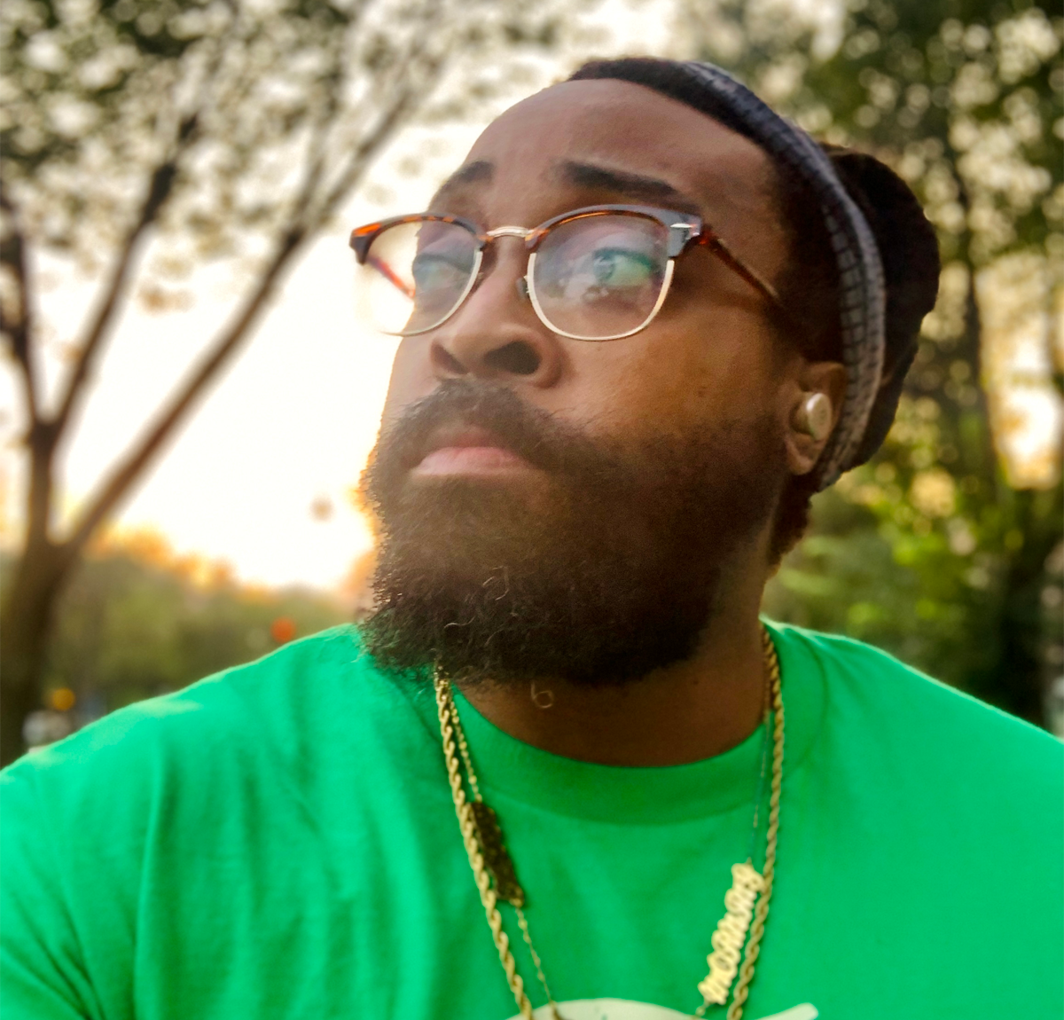 A person wearing a black and grey beanie and glasses with a gold necklace and green shirt.