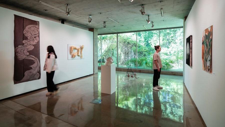 Guests look at student artwork in a bright gallery space bathed in reflective light, a glass wall looking outdoors in the background