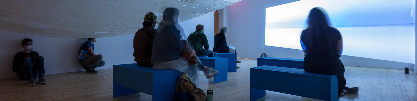 people sitting on blue benches in a room with a projected image on the wall.