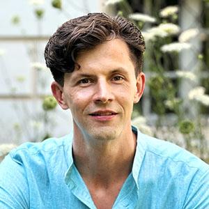 A man with short brown hair wears a turquoise collarless shirt with queen anne's lace out of focus in the background