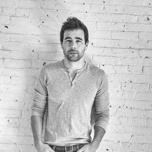 A person with short dark hair and beard wearing a white shirt, standing in front of a white brick wall