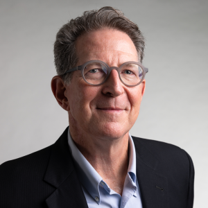 A man with short brown hair and glasses wears a blue button down shirt unbuttoned at the top with a navy blazer