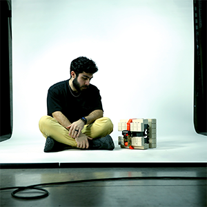 A man sittingon the floor, looking down at a wooden model next to him that is held together with a ratcheting strap.