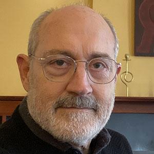 A bearded man with glasses sits indoors against a warm-toned background.