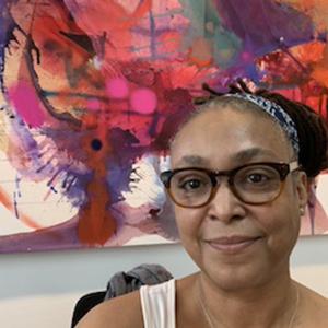 A woman sitting in front of a colorful painting and facing the camera. She has dark hair pulled back from her face and is wearing round glasses.