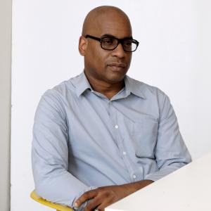 A photo of a dark-skinned bald man in three-quarter profile. He is wearing a light blue button down shirt and black frame glasses and looking off into the distance.