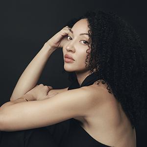 A woman with curly black hair, wearing a black tank top, and resting her head against her hand.