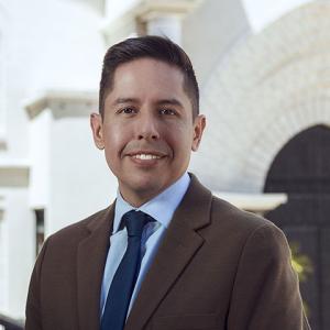 Man with dark, closely trimmed hair wearing a brown suit jacket with light blue shirt and navy tie