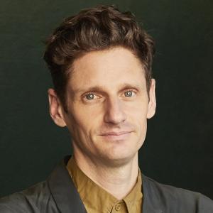 A man with short curly brown hair smiling softly at the camera. There is a dark green background.