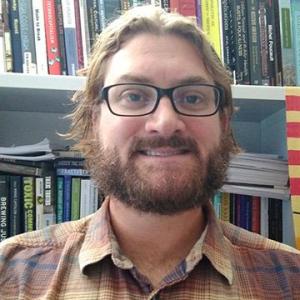 Man with black glasses and ginger colored hair and beard wearing a plaid collared shirt