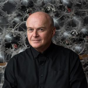 A senior, fair-skinned man with clear eyes wearing a black button-up shirt looking at the camera.