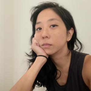 A woman with black hair propping her head up on her hand and looking at the camera