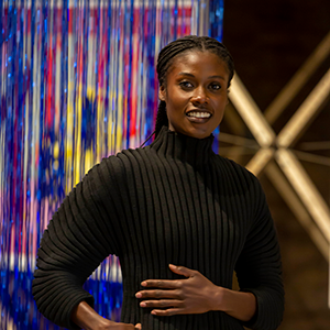 A woman with dark skin wearing a black turtleneck smiles against a colorful textile background.