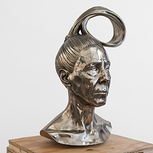 On top of a wood block sits a metal sculpture of a bust with feminine features and hair styled in a loop atop the subject's head, against a white background.