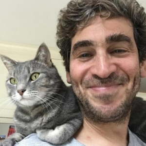 A man with curly brown hair smiling at the camera. There is a grey tabby cat on his shoulder