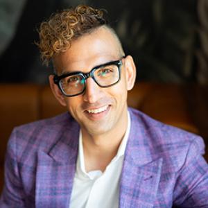 Man smiling with light brown curly hair, wearing thick black framed glasses, a white collared shirt and purple jacket.