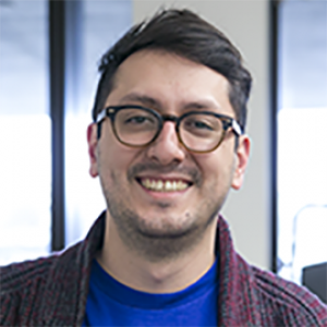 A man with short dark brown hair, wearing round, brown glasses and a blue shirt.