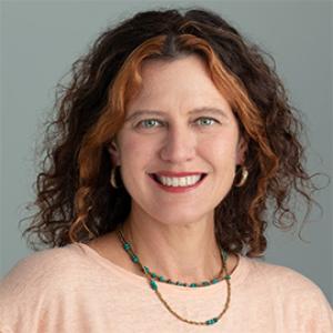 woman smiling with dark brown curly hair and a necklace and pink shirt.