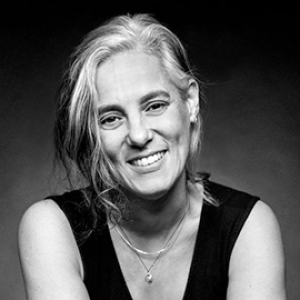 Black and white photo of a pale skinned person with grey hair and feminine features looks directly at the camera, wearing two thin necklaces and a black tank top. 