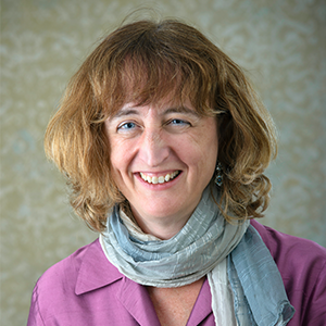 A person with blue scarf and purple blazer smiling in front of a tan backdrop.