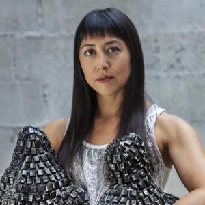 Woman with long dark hair and bangs holding two black sculpture pieces standing in front of a cement wall.
