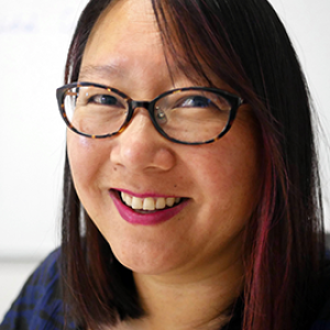 Close up color photograph of a person with feminine facial features and a medium skin tone, wearing pink lipstick, brown eyeglasses, and a navy blue blouse. 