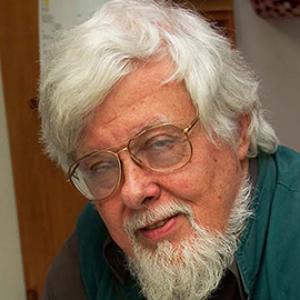 An elderly man with white hair and a beard, who is wearing round wire rimmed glasses and a turquoise shirt. 