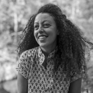 Woman with long dark curly hair wearing a short sleeved blouse with an abstract pattern on it.