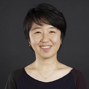 Portrait of a woman with short black hair and wearing a neutral tricolored shirt against a dark grey background