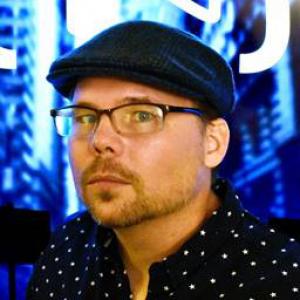 Man wearing glasses, a dark hat and polka dotted shirt against a bright blue hued background.