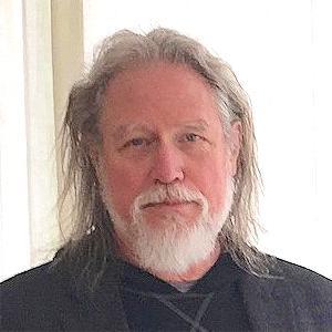 A man with shoulder length white and grey hair, with a short white beard. The man is wearing a black shirt. 