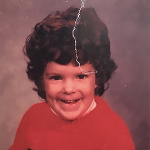 A young child with light skin and dark brown curly hair smiles mischieviously at the camera. 