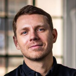 A man with short, light-brown hair and light-colored eyes wearing a black T-shirt.