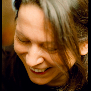 A candid portrait of a woman with long brown hair, smiling and looking down. Her face is partially obscured by her hair, and her expression is warm and relaxed. The soft lighting and close-up focus give the image an intimate and natural feel.