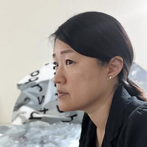 Profile of an Asian woman wearing a black shirt, with dark hair pulled back into a ponytail and stud earrings.