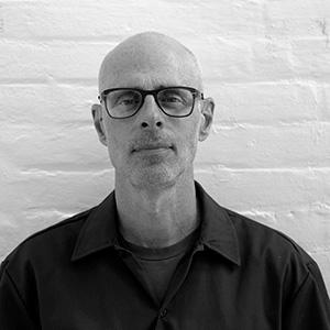 A black and white headshot of a bald man with black glasses and light stubble. He his wearing a dark button-down shirt and standing against a white brick backdrop. 