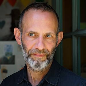 A man with light skin, dark thinning hair, and a salt and pepper beard, wearing a dark blue shirt.