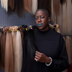 A person with feminine features and closely-shaved dark hair, wearing blue lipstick, a black shirt, and assorted jewelry, stands next to bundles of hair.