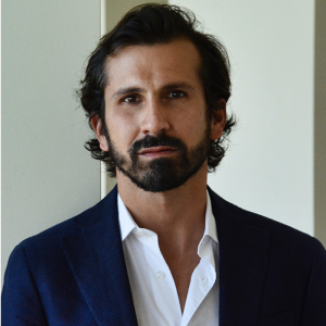 Portrait of a person with brown hair and a beard, wearing a light-colored shirt and a dark-colored jacket.