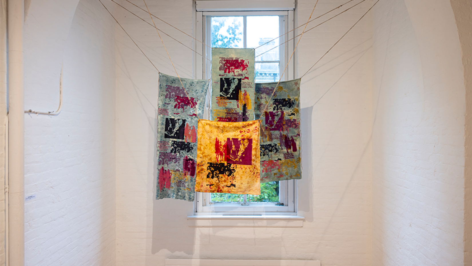 Colorful fabrinc hangs in a white gallery space in front of a window.