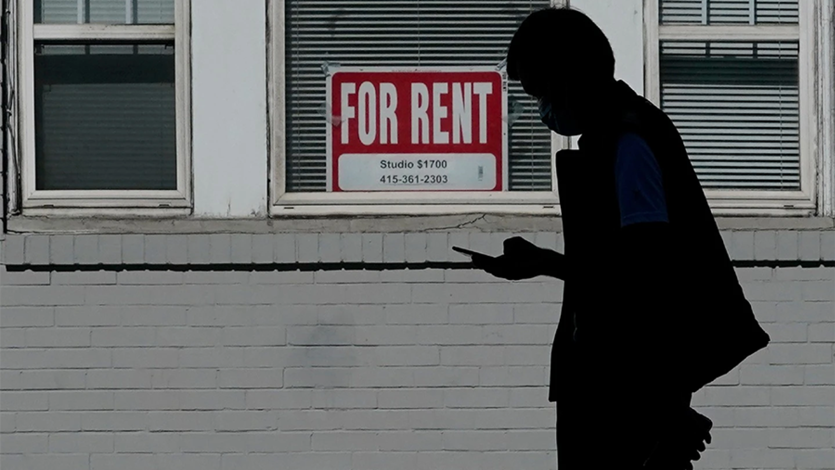 Profile of a person in shadow looking at their cellphone with a grey building in the background displaying a For Rent sign