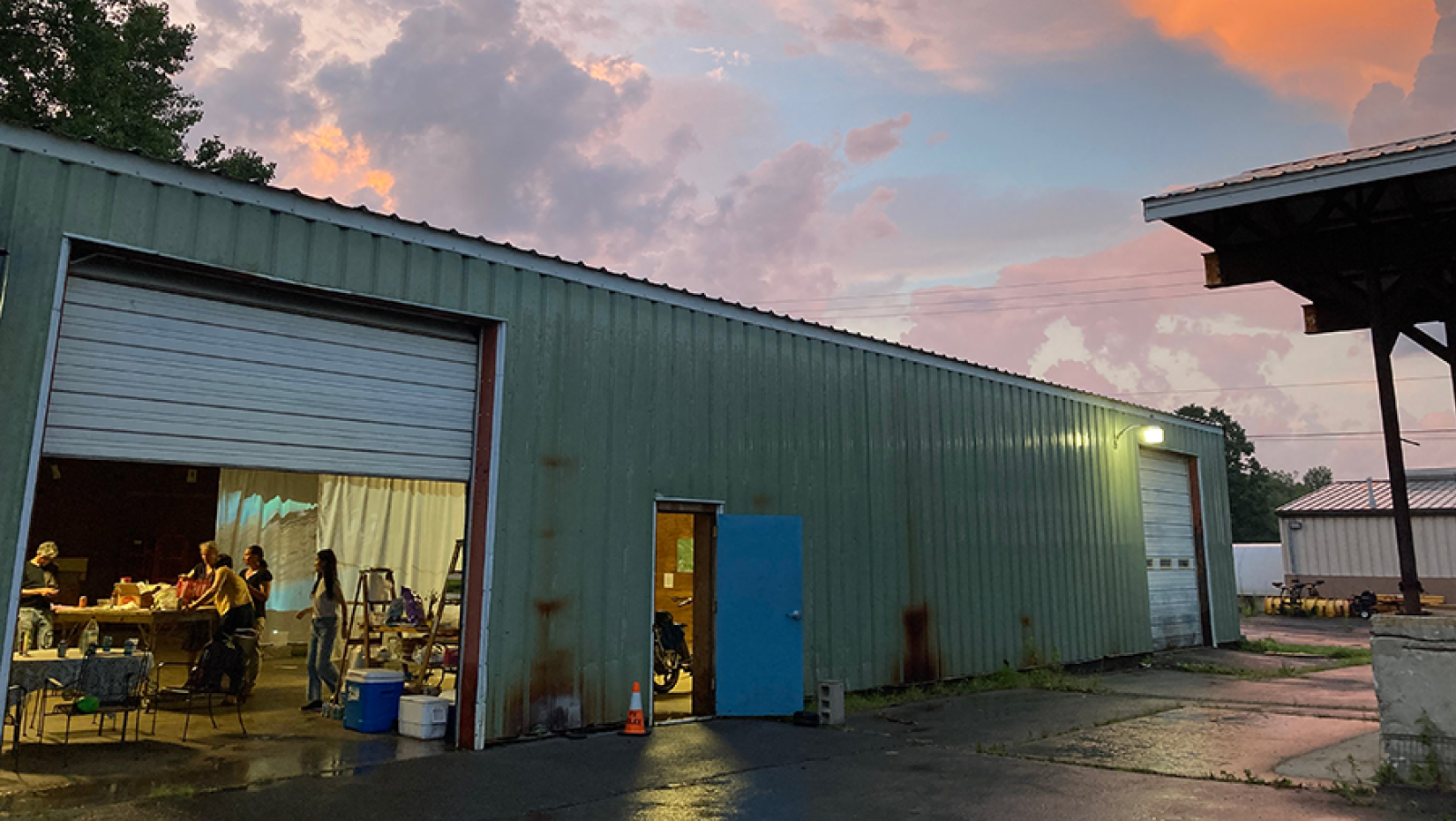 Metal-sided garage space with door rolled up and people inside working at a table