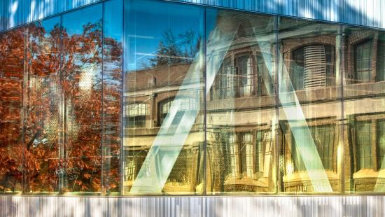 a windowed building corner reflecting the adjacent scenery of fall trees and nearby buildings.