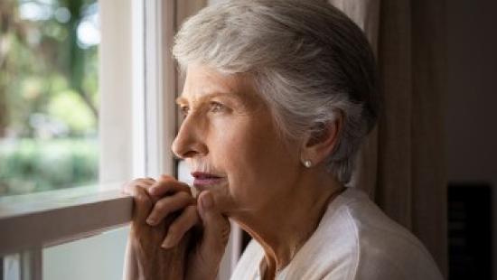 An older woman with short gray hair gazes thoughtfully out a window, resting her hands lightly on the windowsill as soft daylight illuminates her face.