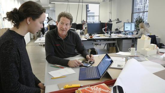 student and faculty member sitting in studio, laptop and books on the desk